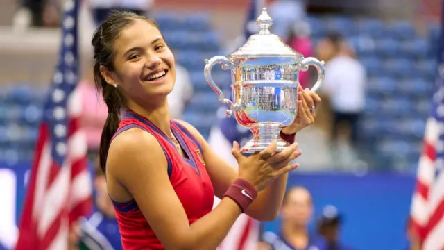 Emma Raducanu, wearing a red sleeveless top and with her dark hair tied back, smiles and poses with the silver US Open trophy held to her left. USA flags and blue stadium seating fills the background