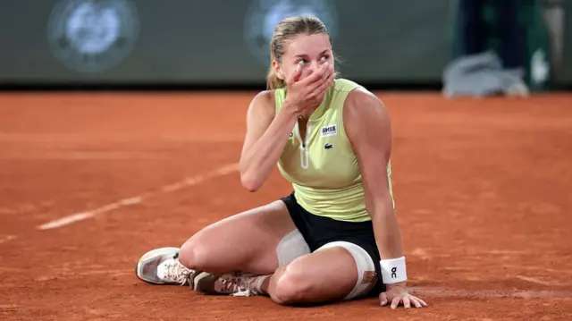 Maja Chwalinska, wearing a yellow sleeveless top and black skirt, sits on the floor with her left hand on the clay and her right hand to her mouth in shock after reaching the final