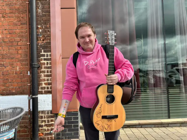 Ayaz, wearing a pink hoodie and holding his signed guitar