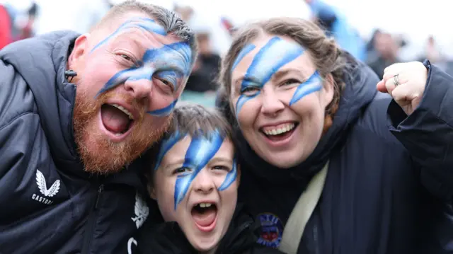 Bath fans - a man, a boy and a woman - wearing blue and white face paint with mouths open