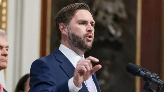 JD Vance is pictured speaking behind a microphone, gesturing with his right hand.