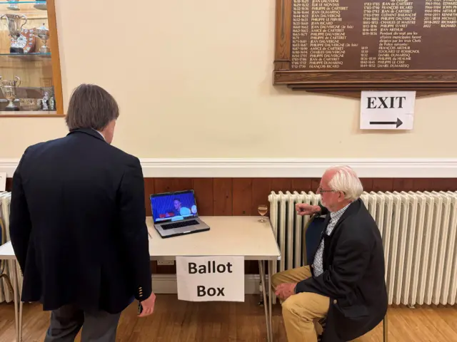 Two men watching a screen. They are sat together next to a white table which has a sign saying Ballot Box.