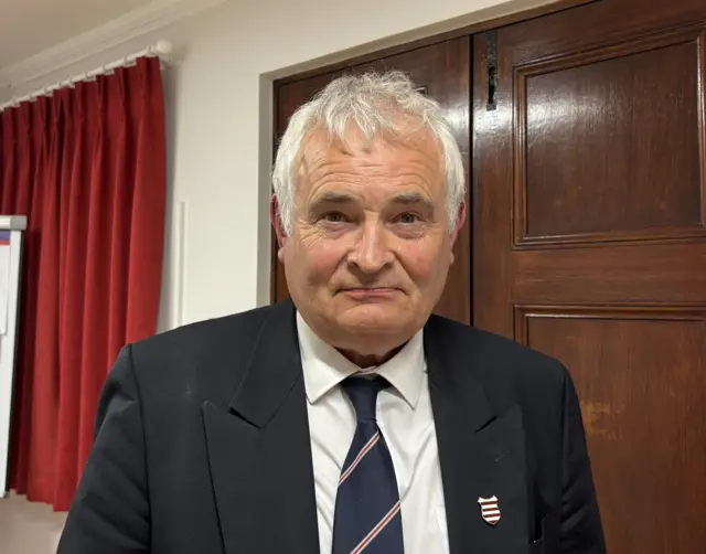 A picture of Mark Labey - he is wearing a white suit shirt. a navy tie and a black blazer.