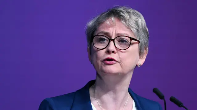 Foreign Secretary Yvette Cooper wearing a navy blazer while standing in front of a microphone. A purple background is behind her.