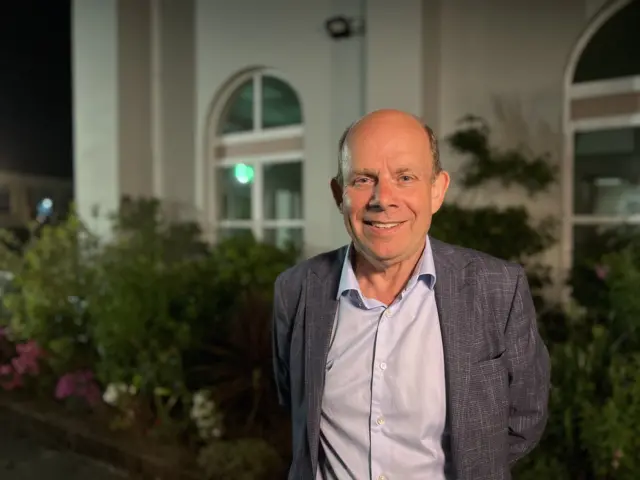 A man wearing a blue t-shirt and dark grey blazer. He is stood outside a white building with green bushes.