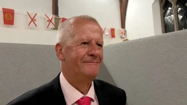 A man wearing a suit and red tie. He is stood in front of a grey wall. In the background there are Jersey flags.
