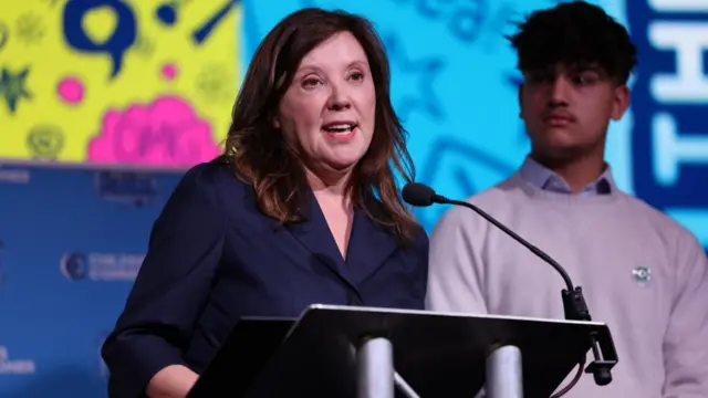 A photo of Rachel de Souza speaking at a podium. Behind her are bright graphics