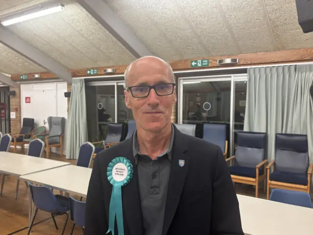 A picture of Jonathan Renouf wearing a grey shirt with a black blazer. He has a blue badge on his blazer and is stood in a large room with tables and chairs.