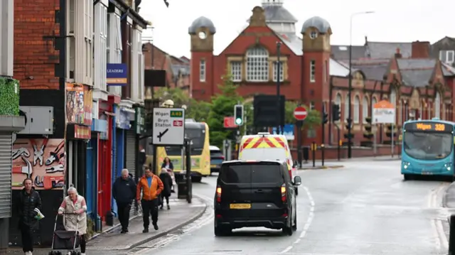 People walk through the town of Ashton-in-Makerfield in Wigan whith vehicles including a turquoise single decker bus heading for Wigan travelling on the road. It is