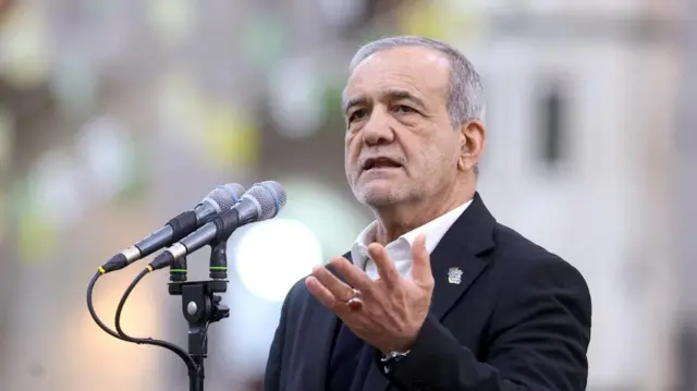 Iranian President Masoud Pezeshkian holds up his hand while giving a speech. He is wearing a dark suit and talking into two microphones.