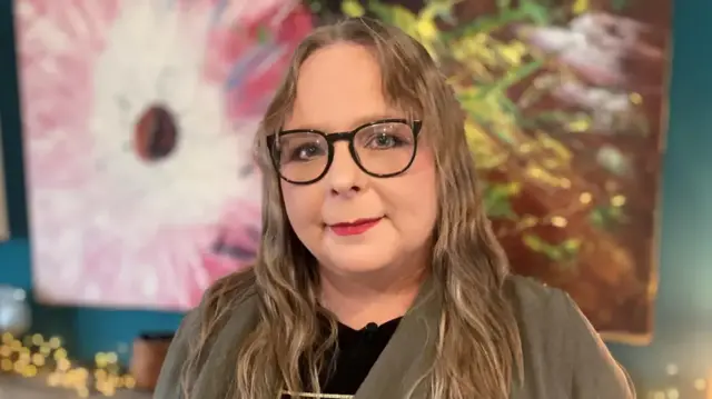Karen Bentham, with long hair and glasses, is sat against a floral background