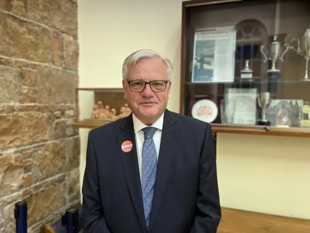 A man wearing glasses, a navy blue blazer, blue tie and white shirt. He is stood in a room with cream and stone walls.