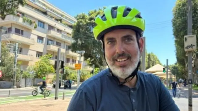 A headshot of Eyal who looks to the camera with a yellow bike helmet on