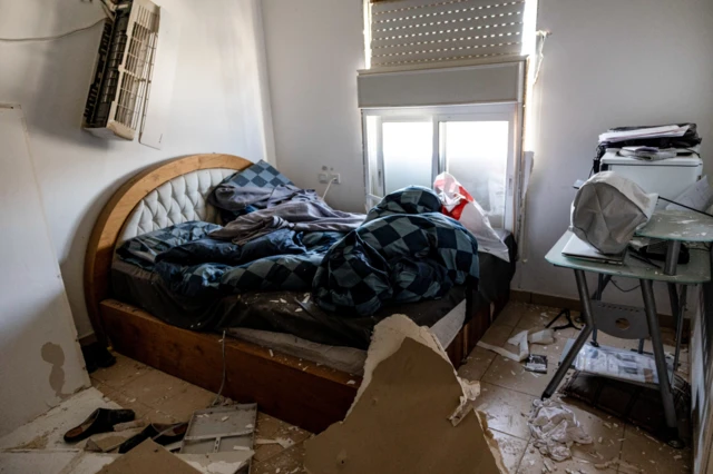 A wooden bed sits in the middle of a room which has been covered in debris and parts of plasterboard from the ceiling, while an electrical fan has been knocked out of place on the wall