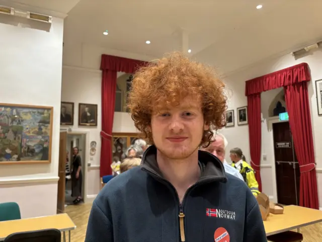 A man with ginger hair. He is wearing a navy blue fleece. Behind him are tables and people in the background.
