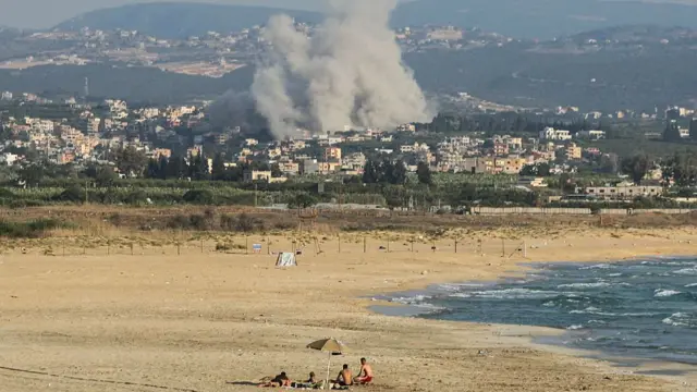 A group sunvathes the beach in the southern Lebanese city of Tyre as smoke rises from the site of an Israeli airstrike in the village of Deir Qanoun Ras al-Ain