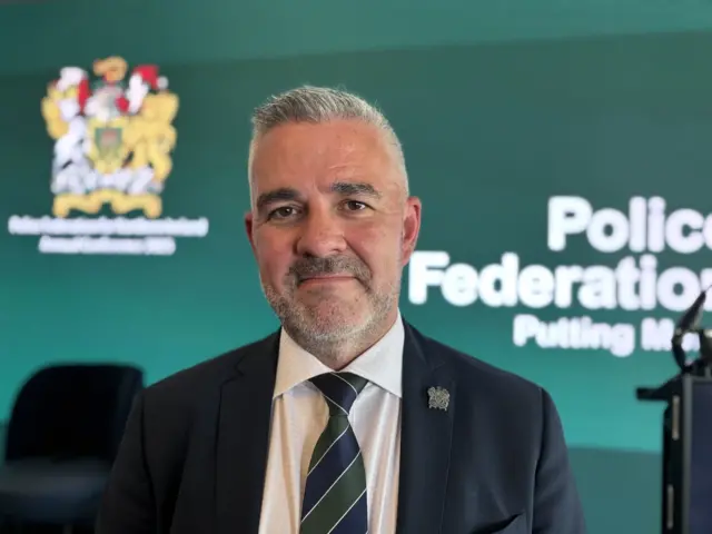 Liam Kelly standing in front of a green background. He is wearing a dark suit and a green and navy tie.