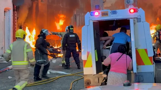 A terraced street where at least two homes are on fire, there are several firefighters on the street, and a hose is strewn across the road. To the right there is a police land rover, with the back doors open where a family - a boy and a mother - have their back to the camera and are entering the vehicle