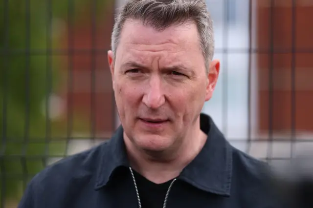 A man in close up, he has short greying hair. He is wearing a blue zip-up jacket and black t-shirt. He is standing outside in front of a metal fence.