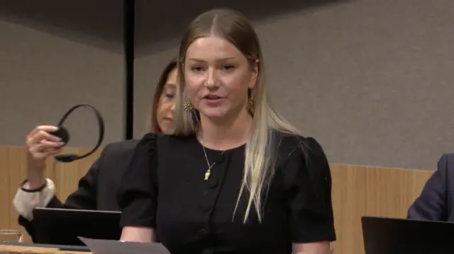 A young woman with blonde hair stands up to speak in the Senedd