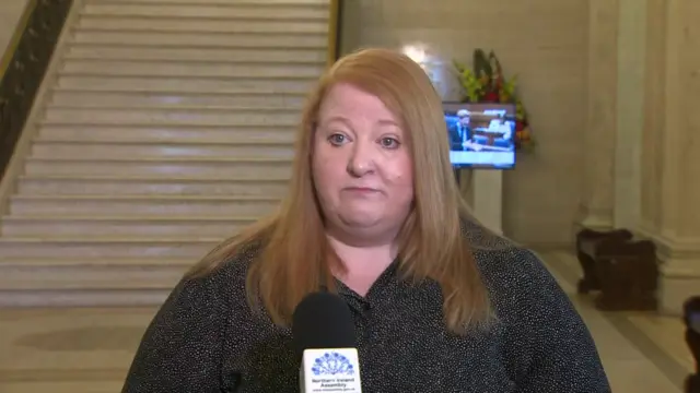 Naomi Long, a woman with long, straight ginger hair, looks at the camera during a Stormont press conference.   She is wearing a black blouse with small white dots.