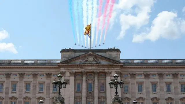 The Red Arrows aircraft are shown flying over Buckingham Palace, leaving a trail of red, white and blue smoke