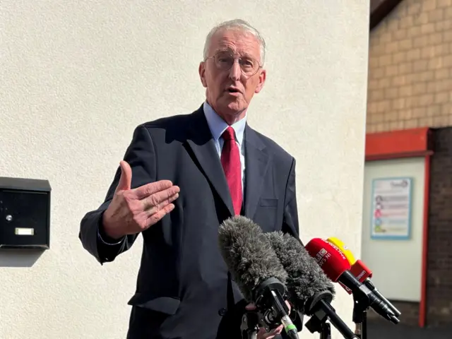 Hilary Ben standing before mics- black suit, red tie, grey hair and glasses