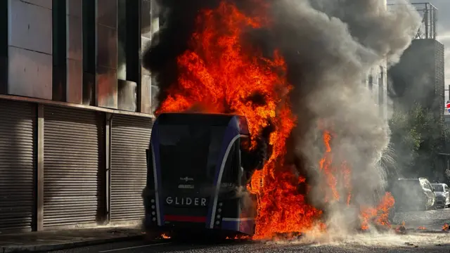 Flames engulf a bus on a city street. The bus, with purple stripes to the rear, says Glider across the back. The flames are across the middle of the vehicle. In the background is a building with shutters down.