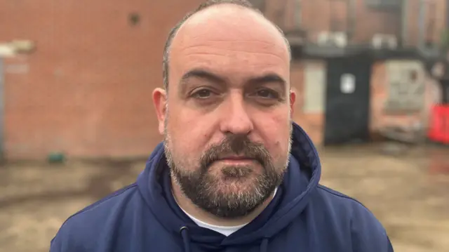Paul McCusker, mid shot, black hair with beard wearing blue hoody. In front of a red brick building