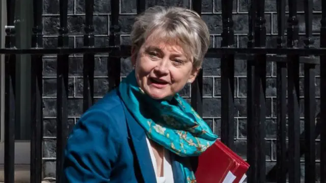 Yvette Cooper, wearing a blue suit and turquoise scarf, walks along the pavement into Downing Street while carrying a red folder and turning her head towards the camera