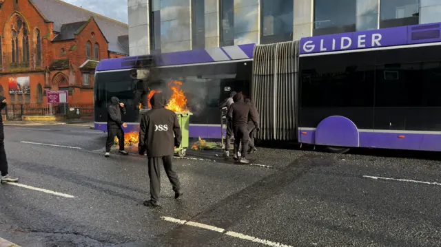 A glider bus being attacked by masked people and a bin is on fire.
