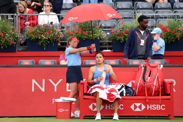 Emma Raducanu of Great Britain is sheltered by an umbrella held up by a ball kid as rain delays play during the Women's Singles first round match against Anna Blinkova