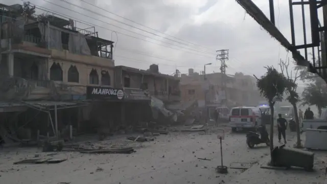 A building has been destroyed and rubble is strewn on a road, three ambulances can be seen next to it