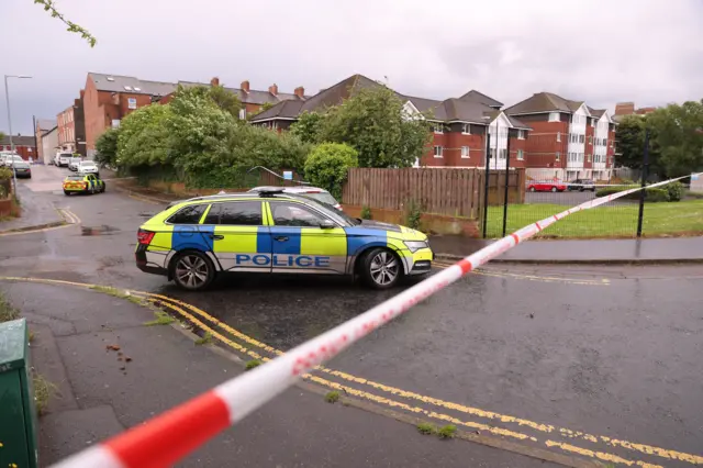 A police car parked behind a cordon at the scene of a serious stabbing incident on Kinnaird Avenue in north Belfast. A block of apartments and red brick houses are in the background.