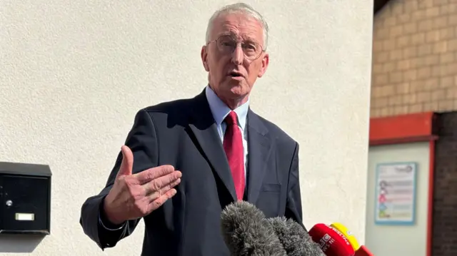 Hilary Benn Secretary of State standing in front of microphones. Black suit, blue shirt and red tie. He has grey hair and is wearing glasses