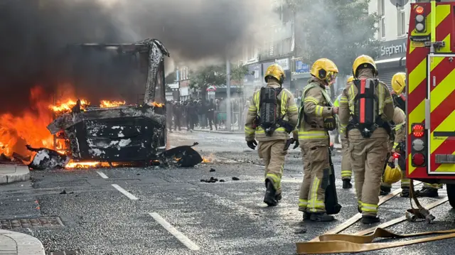 Fire fighters at a fire engine attend to a bus on fire - the bus is mostly destroyed, with flames still seen inside the husk. In the background a large group of people can be seen