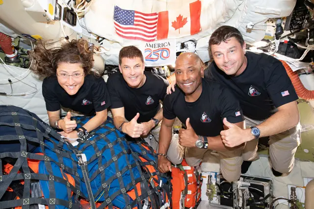 The Artemis II crew pose for a photo with the US and Canadian flags behind them. Also behind them is a flag with "America 250".