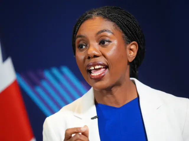 Kemi Badenoch standing in front of a dark blue background. She is wearing a blue shirt and a white jacket.