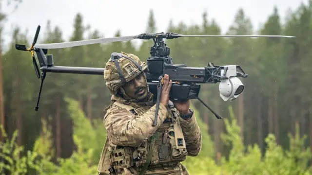 A soldier dressed in camo carries a Ghost drone. The drone has a rotor and is styled like a helicopter. It has a camera attached to the end of it