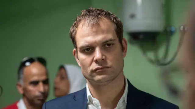 Hamish Falconer wears a navy suit and stands in front of a green wall background. A man and a woman can be seen behind him.