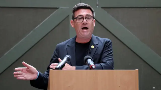 Andy Burnham gestures as he stands at a wooden podium while delivering a speech in Manchester