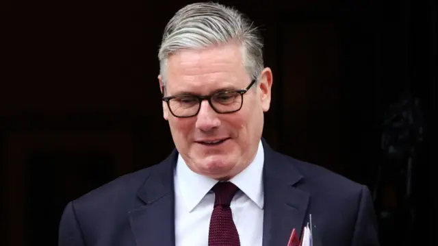 Keir Starmer looks at the ground as he departs 10 Downing Street. He is wearing glasses, a blue suit, white shirt and red tie