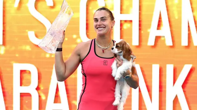 Sabalenka, wearing a pink dress, smiles and holds a glass trophy in her right hand and her brown and white spaniel Ash under her left arm
