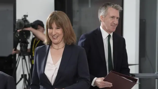 Rachel Reeves smiles as she walks across a room. Dan Jarvis can be seen behind her holding a burgundy folder