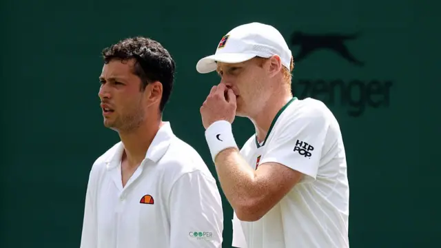 Joshua Paris (left), wearing a white polo shirt, looks down court open mouthed, while Ben Jones (right), wearing a white t-shirt and cap, talks tactics with his left hand covering his mouth