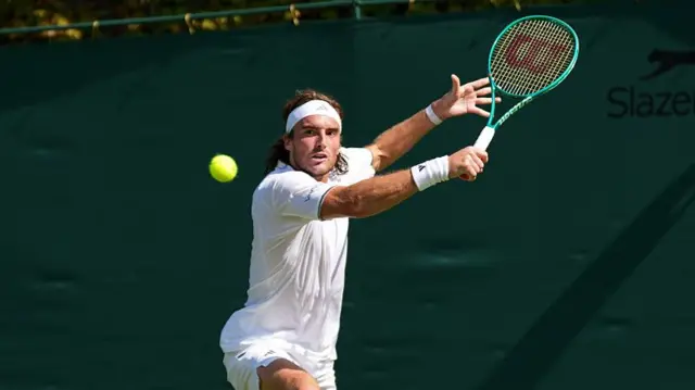 Stefanos Tsitstpas, wearing white t-shirt, shorts and a white bandana over long brown-blonde hair, lunges for a backhand with his green racquet held up in his right hand