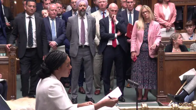 Kemi Badenoch speaking in the House of Commons, with several MPs standing to the side of her.