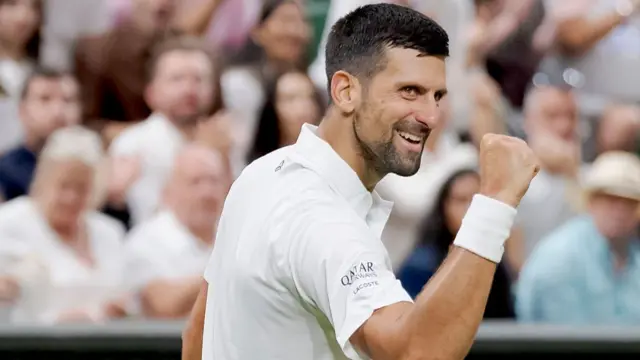 Novak Djokovic, wearing white, smiles with his right fist clenched in celebration