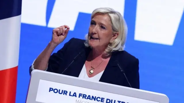 Marine Le Pen is pictured speaking behind a podium in front of a French flag, gesturing with her right hand.