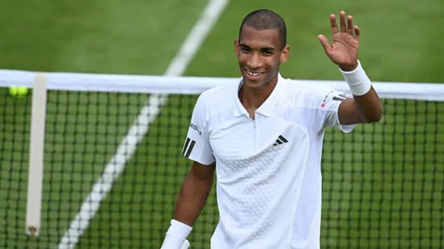 Felix Auger-Aliassime, wearing a white t-shirt, waves his left hand above his head